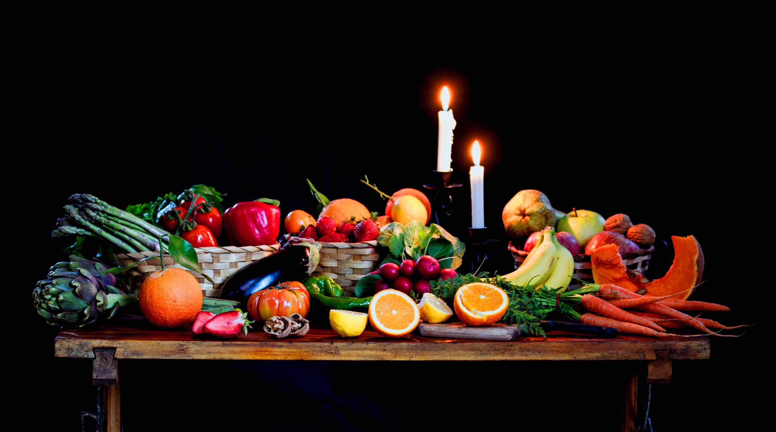 a table sits in a black room, on it there is a pair of bright burning canddles surrounded in a bounty of fruits and vegtables