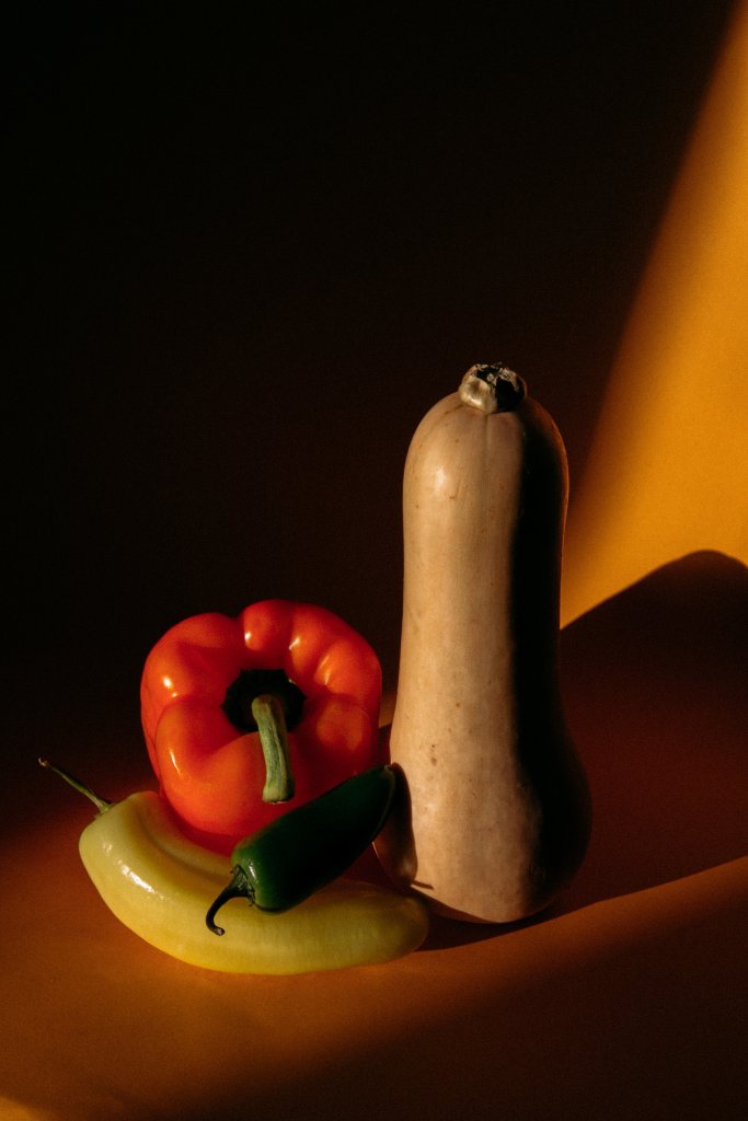 three different peppers and a squash are arranged carfully on a orange table top with strong shaddows cutting across the yellow wall behind them