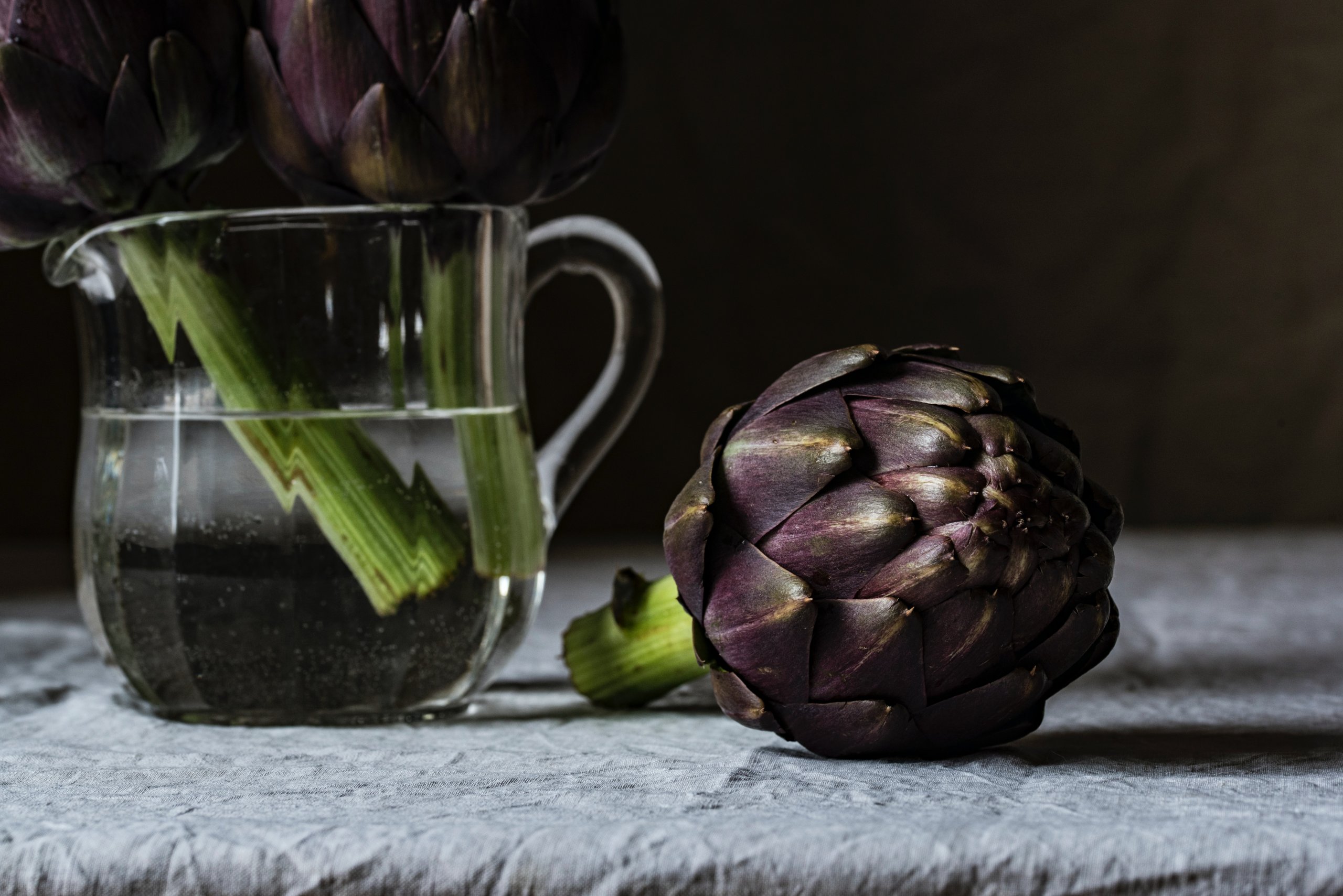 set against a black background, two dark purple heads of artichoke sit in a clear glass jug of water, a third lies on the grey table cloth next to them.