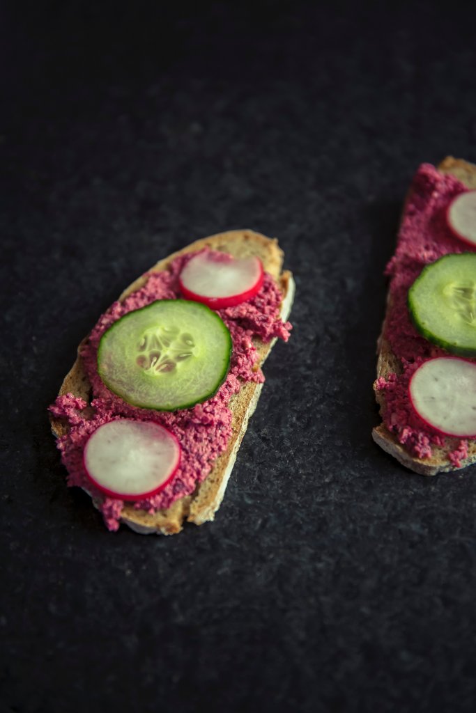 On a black table lies two pieces of sour dough toast with beetroot humus, two pieces of raddish and a slice of cucumber in the middle